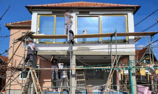 Skela na prednjoj strani kuće sa dva radnika na dnu i jednim na njenom drugom nivou koji izvode radove